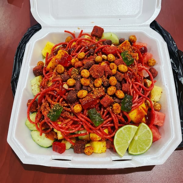 A vibrant Mexican fruit salad with red candy, peanuts, and lime in a white to-go container.