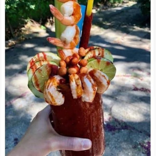 Hand holding a michelada garnished with shrimp, cucumber, lime, and peanuts.