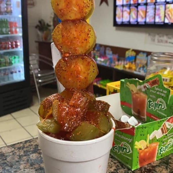 A white cup filled with pickles and a skewer of four yellow fruits, all covered in red seasoning.