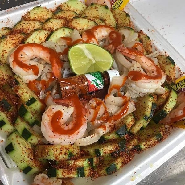 Shrimp and cucumber salad with lime, hot sauce, and chili powder in a white container.