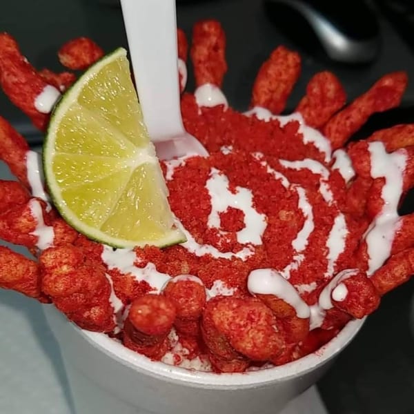 Close-up of a cup filled with red spicy snacks, drizzled with white sauce and a lime wedge.