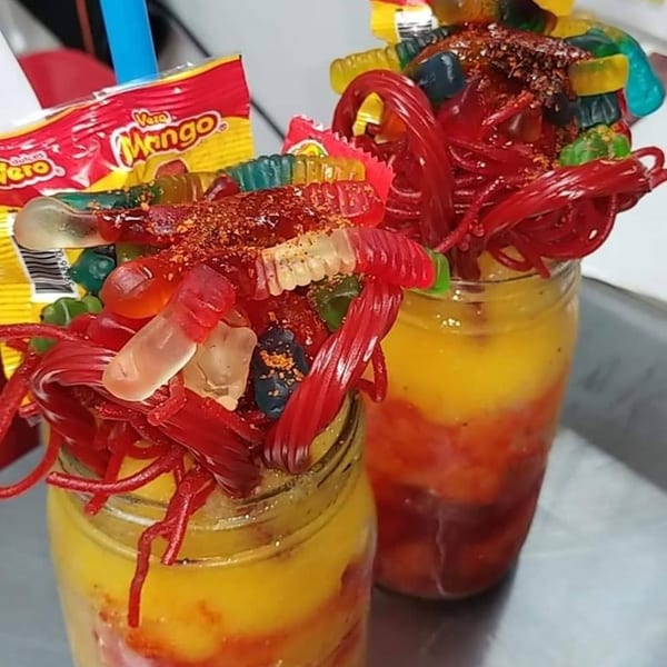 Two vibrant mango drinks in mason jars, topped with gummy candies, red licorice, and chili powder.