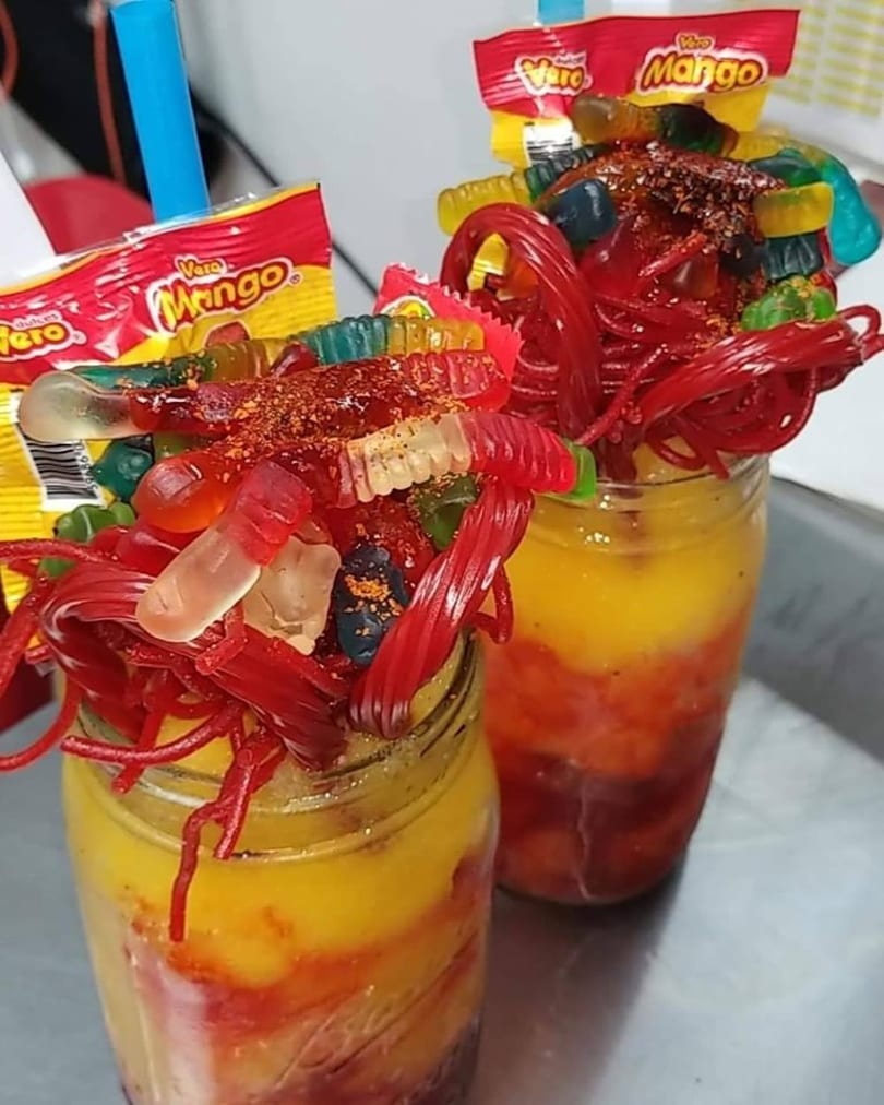 Two vibrant mango drinks in mason jars, topped with gummy candies, red licorice, and chili powder.