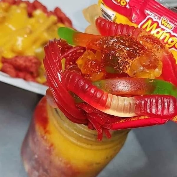 A jar of yellow and red liquid topped with gummy worms, candy, and a Vero Mango candy wrapper.