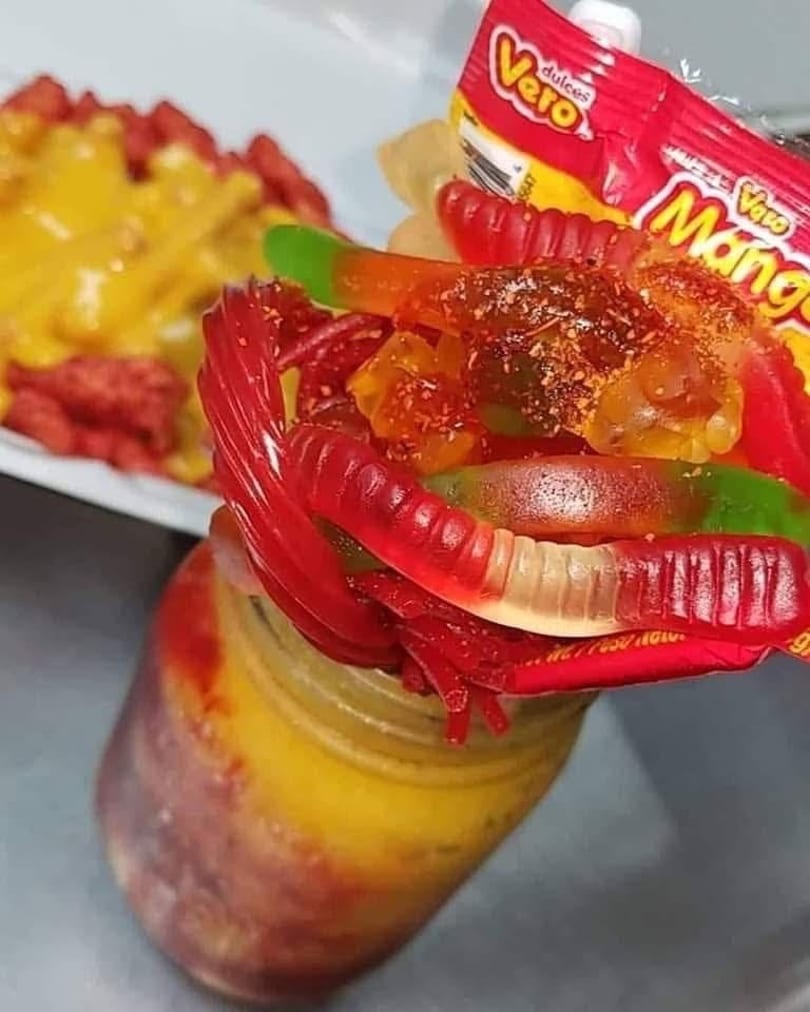 A jar of yellow and red liquid topped with gummy worms, candy, and a Vero Mango candy wrapper.
