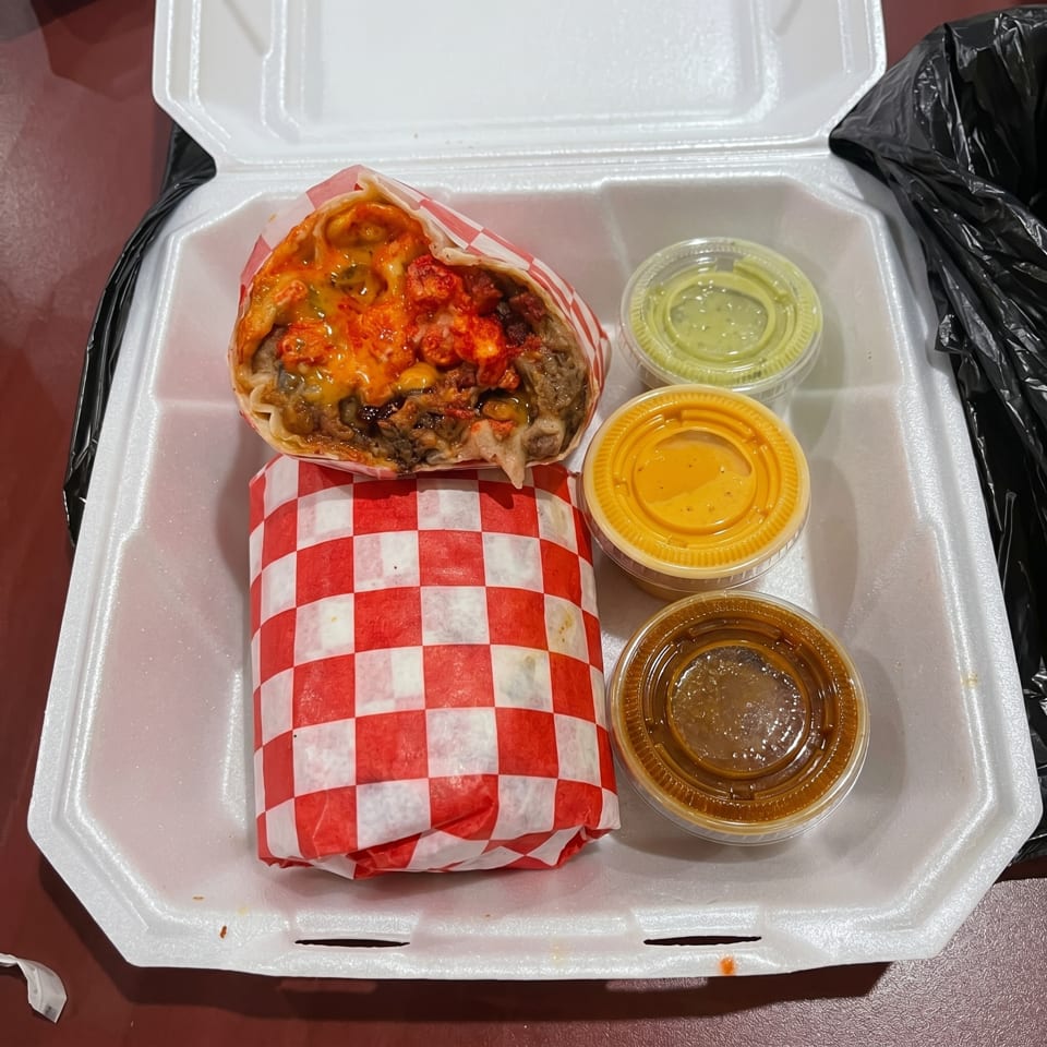 A takeout container with a burrito, one wrapped in red and white paper, and three sauce cups.