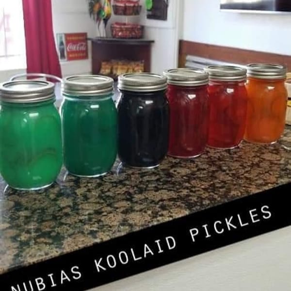 Six colorful jars of Kool-Aid pickles, including green, black, red, and orange, on a speckled countertop.