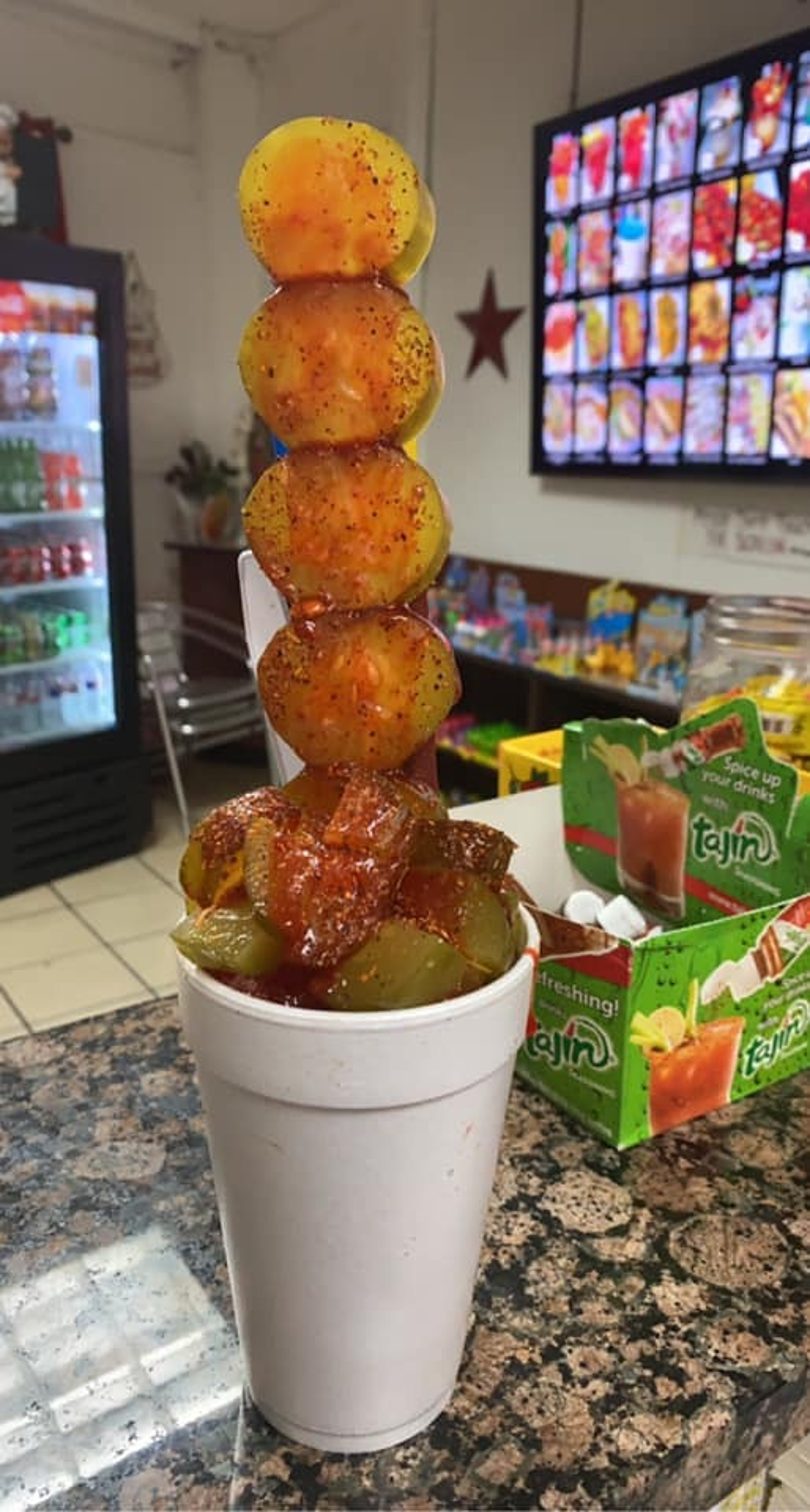 A white cup filled with pickles and a skewer of four yellow fruits, all covered in red seasoning.