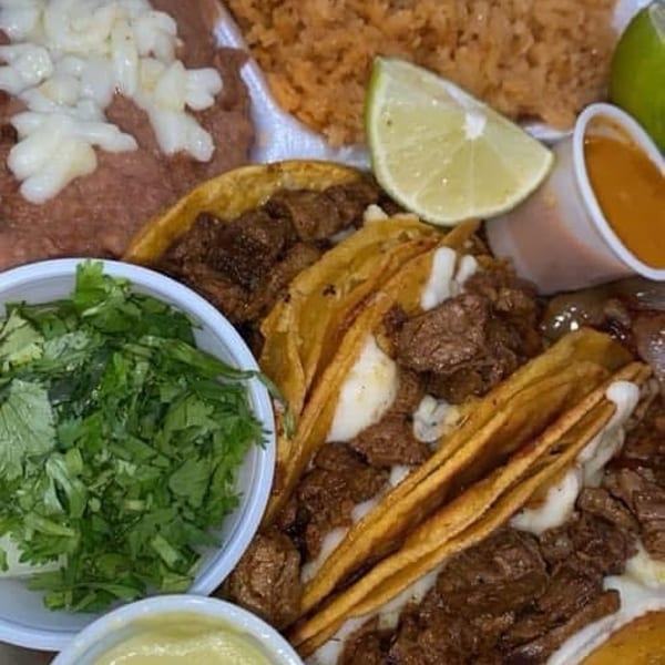 A takeout container holds steak tacos with cheese, rice, refried beans, cilantro, and sauces.