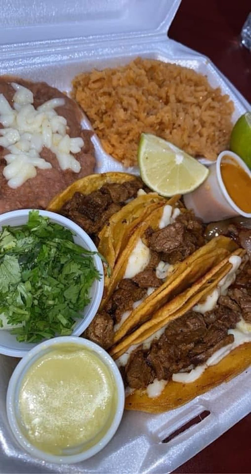 A takeout container holds steak tacos with cheese, rice, refried beans, cilantro, and sauces.