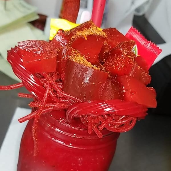 A vibrant red drink in a jar, topped with red gummy candies, licorice, and chili powder.