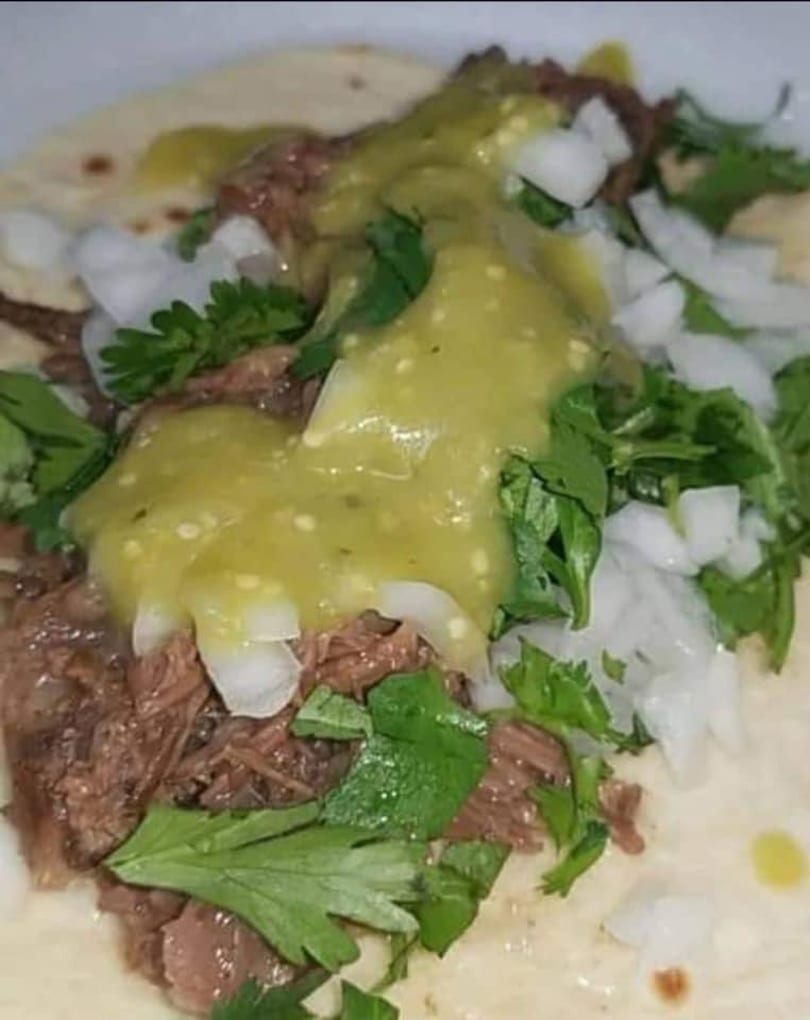 Close-up of a taco filled with shredded meat, topped with green salsa, diced onions, and cilantro.
