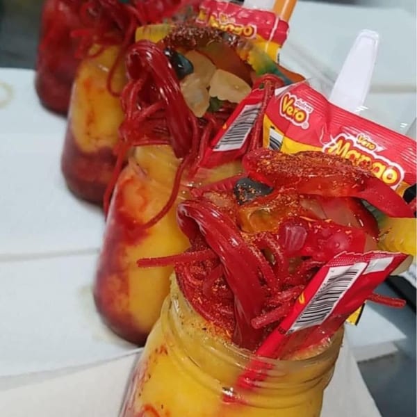 Three vibrant mango drinks in mason jars, topped with red candy, gummies, and chili powder.