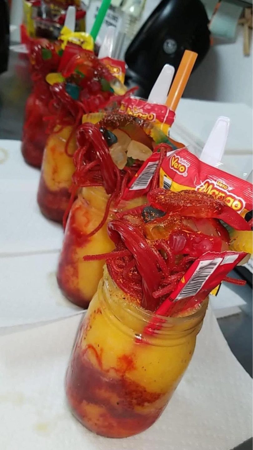Three vibrant mango drinks in mason jars, topped with red candy, gummies, and chili powder.